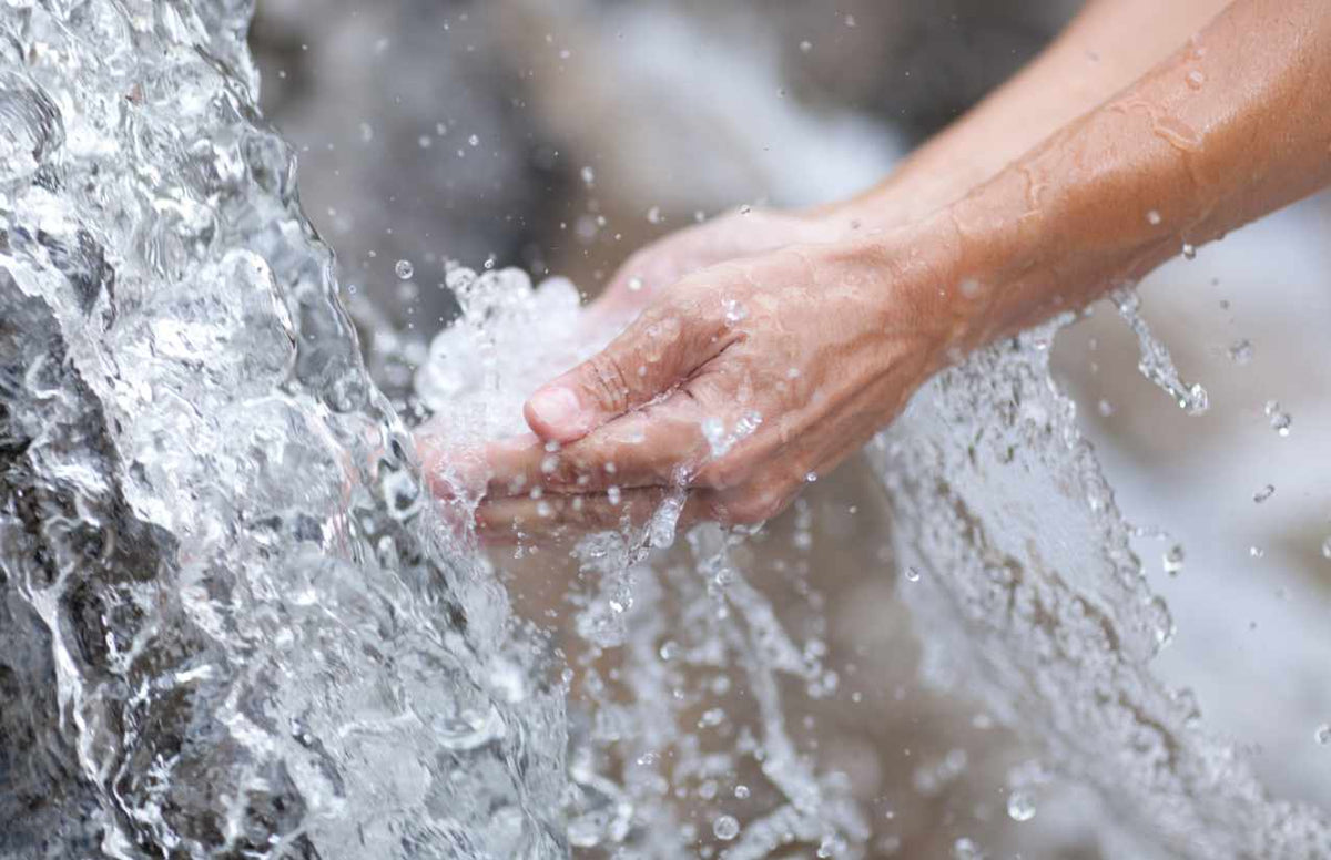 Hände schöpfen sprudelndes Quellwasser aus einem strömenden Bach, Spritzer in der Luft