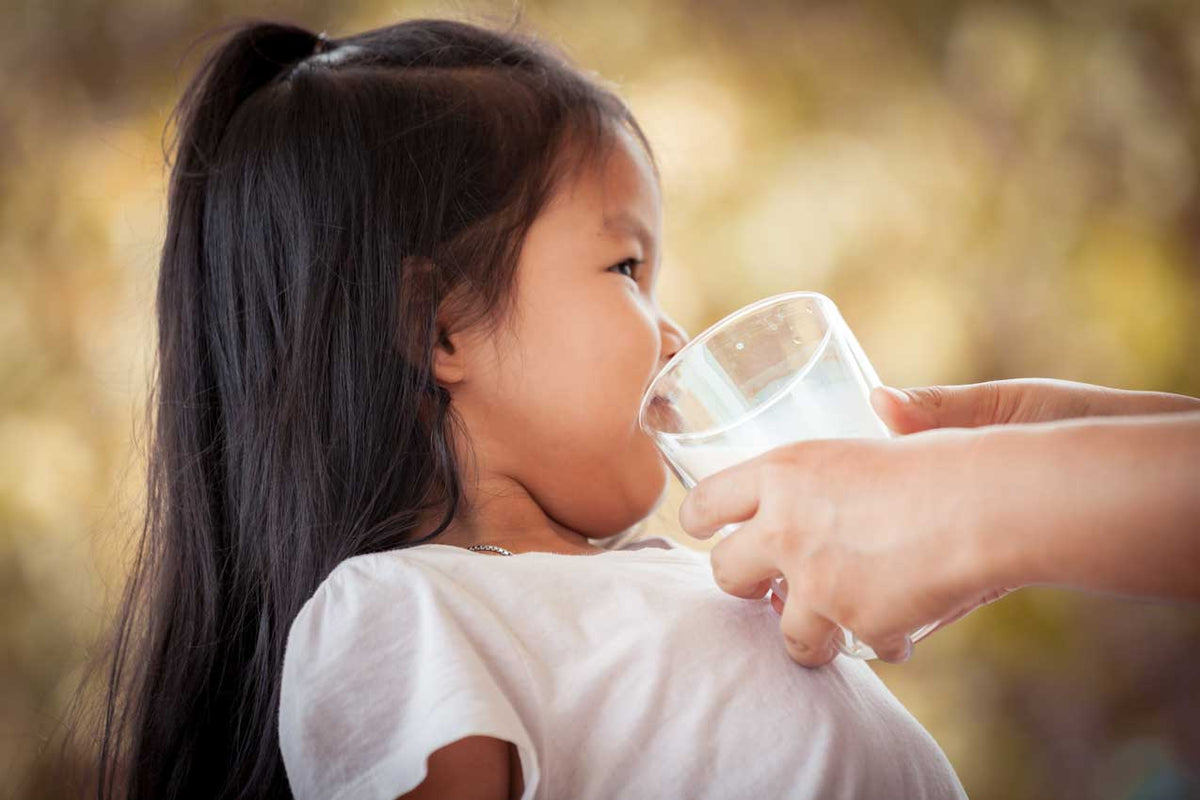 Kleines Mädchen trinkt aus einem Wasserglas, das eine Erwachsenenhand ihm hinhält