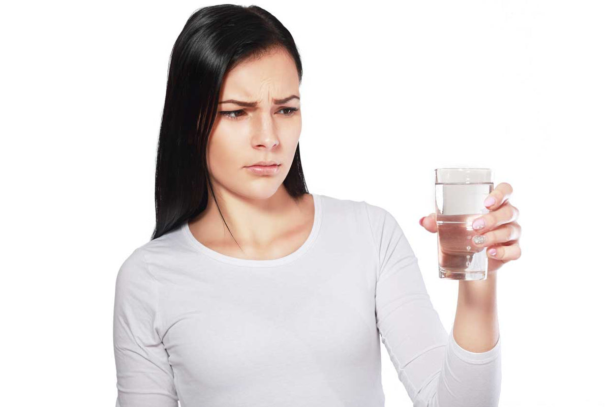 Frau betrachtet skeptisch ein hochgehaltenes Glas Wasser vor weißem Hintergrund