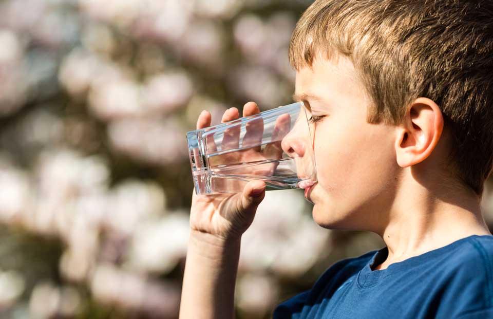 Junge trinkt draußen vor blühendem Baum ein Glas klares Wasser