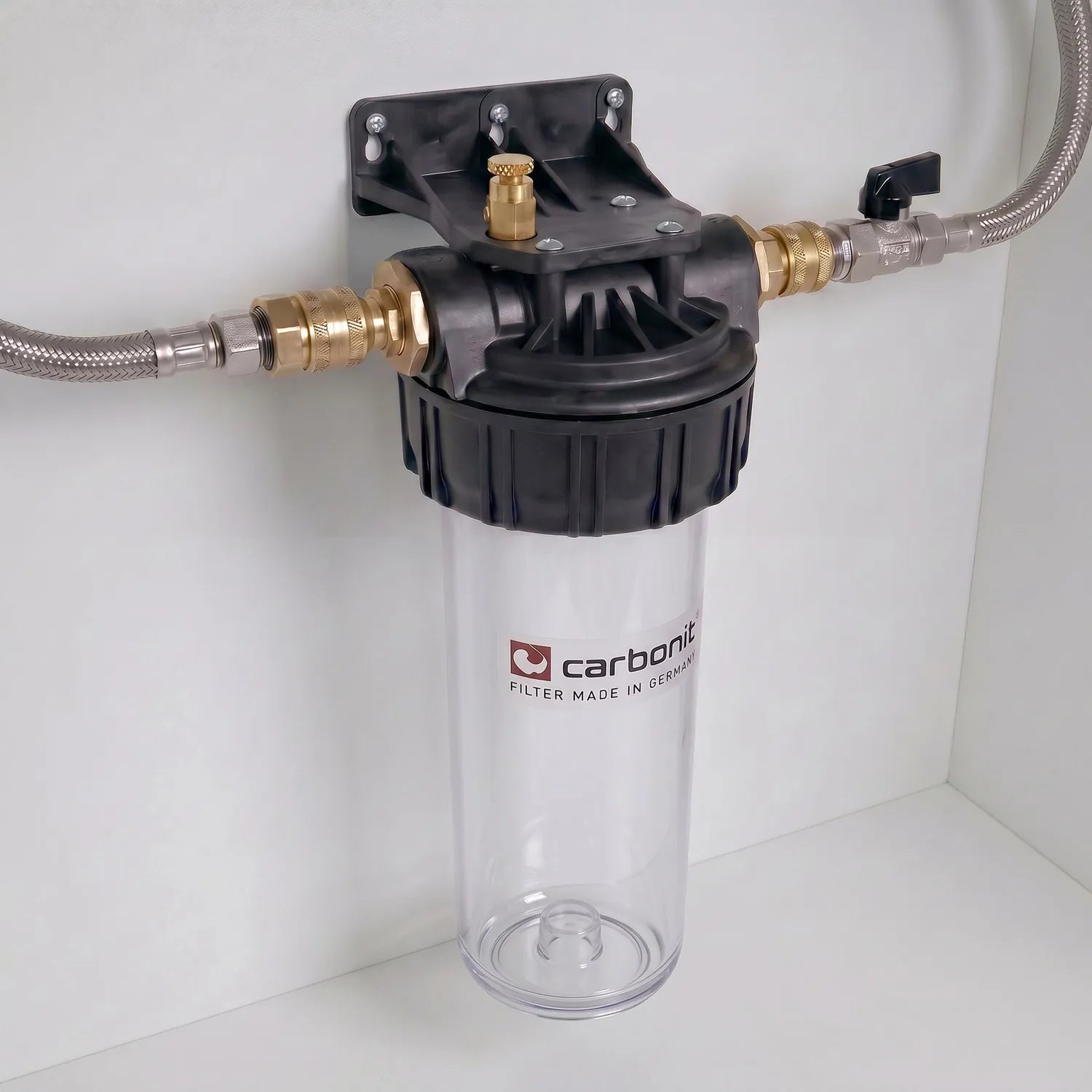 Carbonit Vario Basic montiert an weißer Schrankwand – transparentes Gehäuse mit weißer Filterkartusche (Kreuzstruktur, grüner Ring), schwarzer Kopf mit Messing-Entlüftungsschraube und Schnellkupplungen, zwei Edelstahlschläuche (einer mit schwarzem Kugelventil-Hebel)