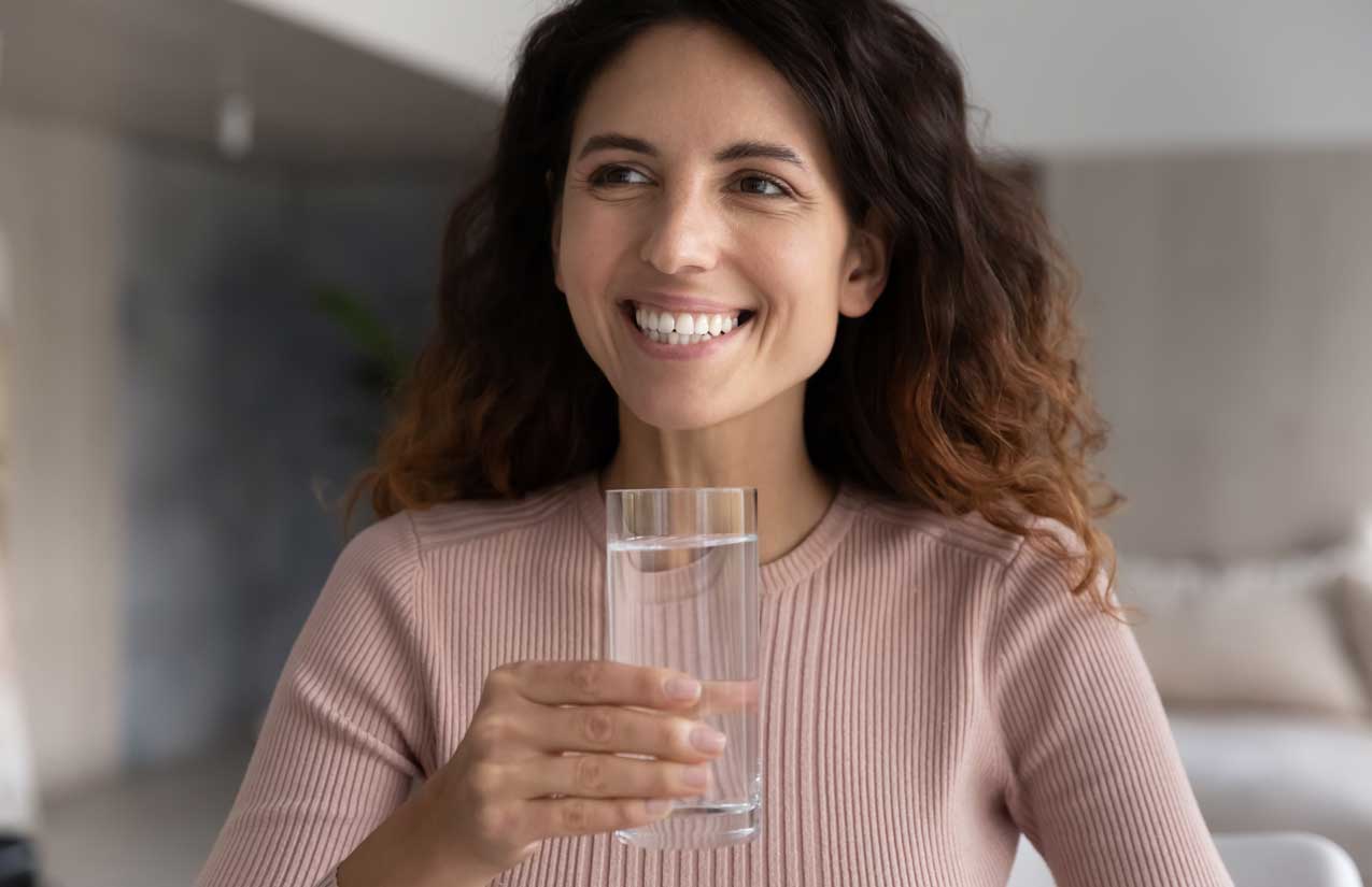 Lächelnde Frau hält im Wohnraum ein Glas mit klarem Wasser in der Hand