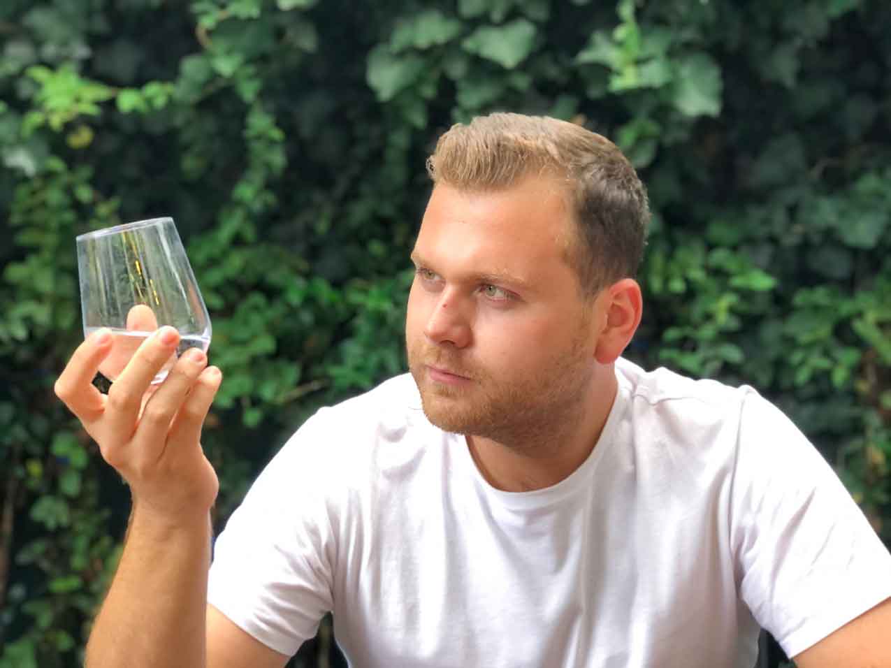 Mann im weißen Shirt hält ein Wasserglas gegen das Licht und prüft dessen Färbung