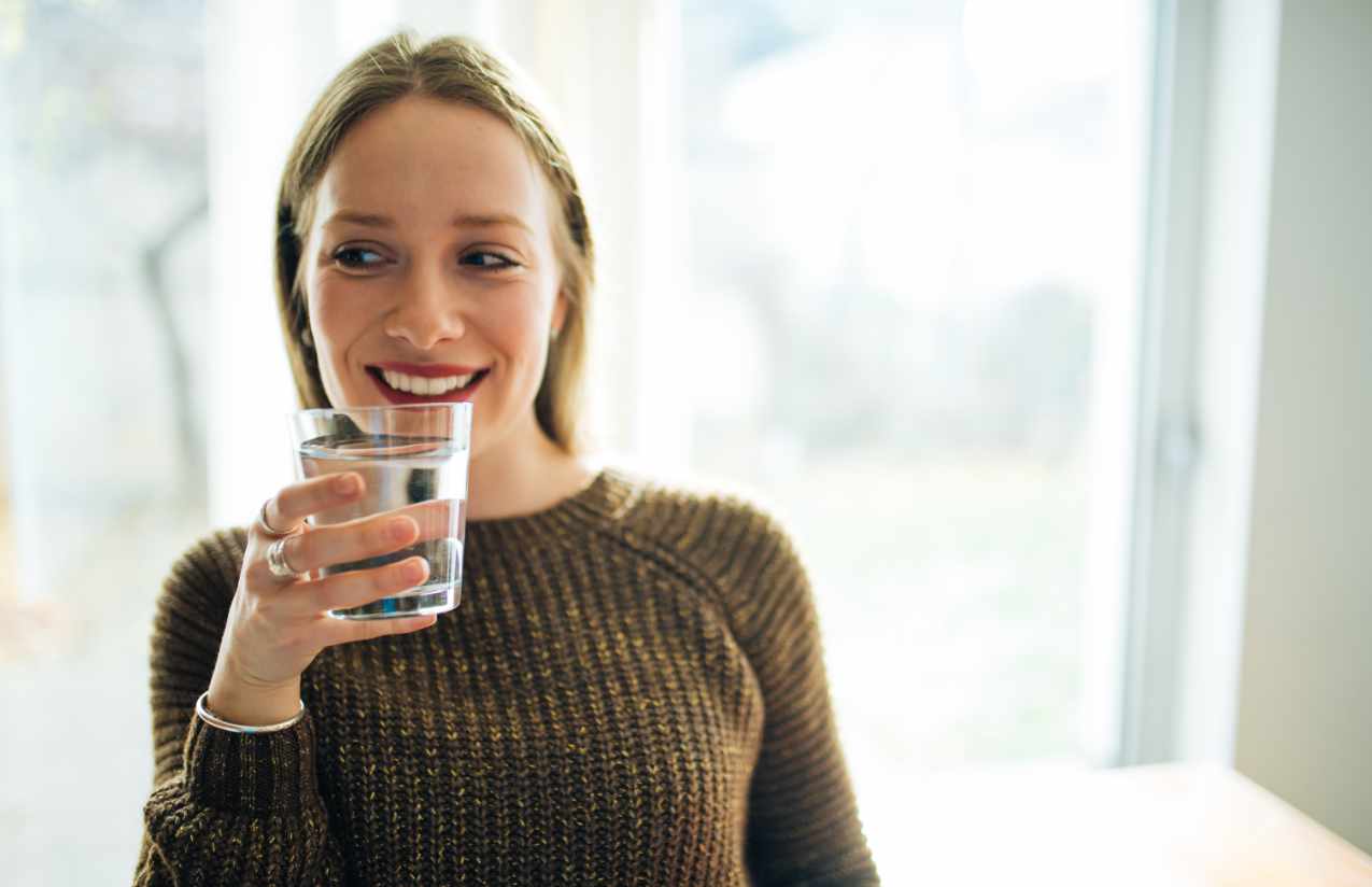 Lächelnde junge Frau am Fenster hält ein Glas klares Trinkwasser in der Hand