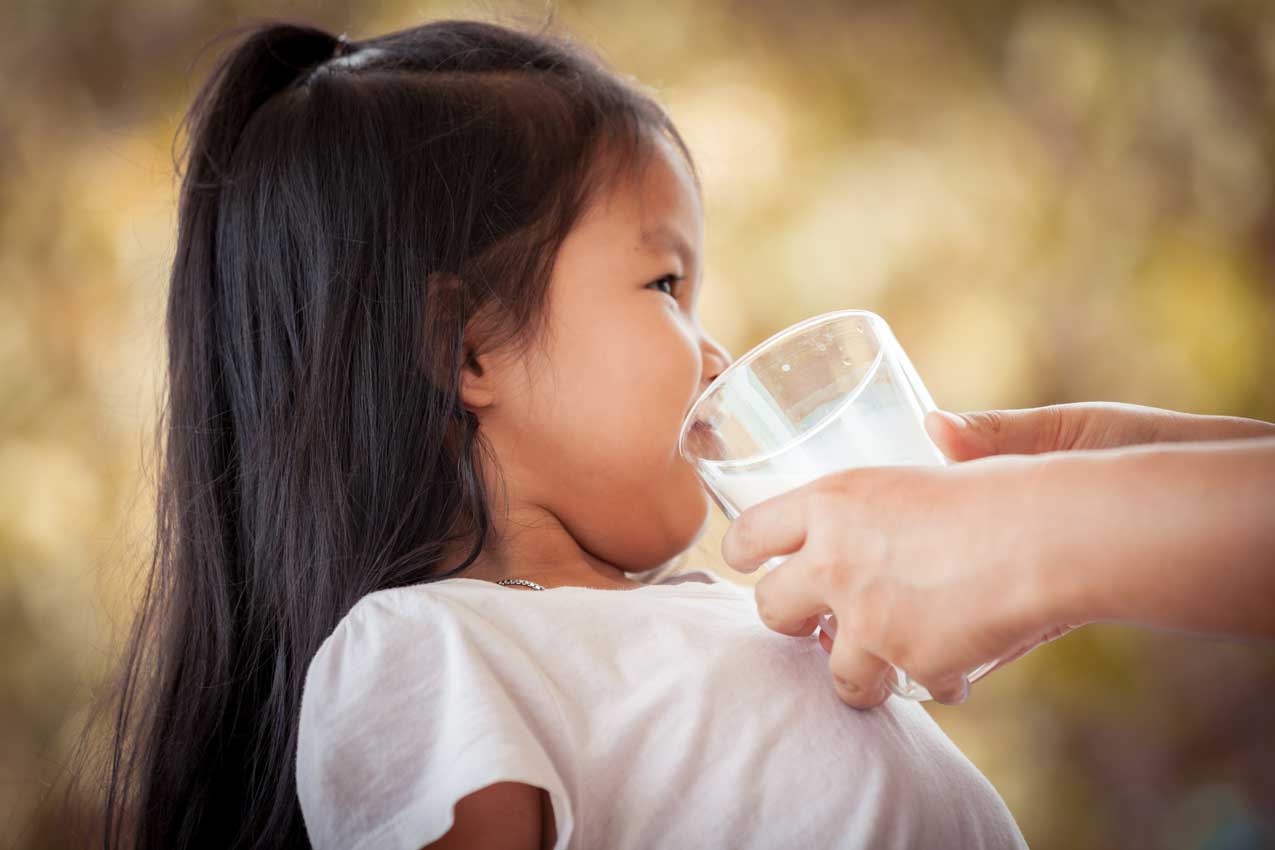Kleines Mädchen trinkt aus einem Wasserglas, das eine Erwachsenenhand ihm hinhält