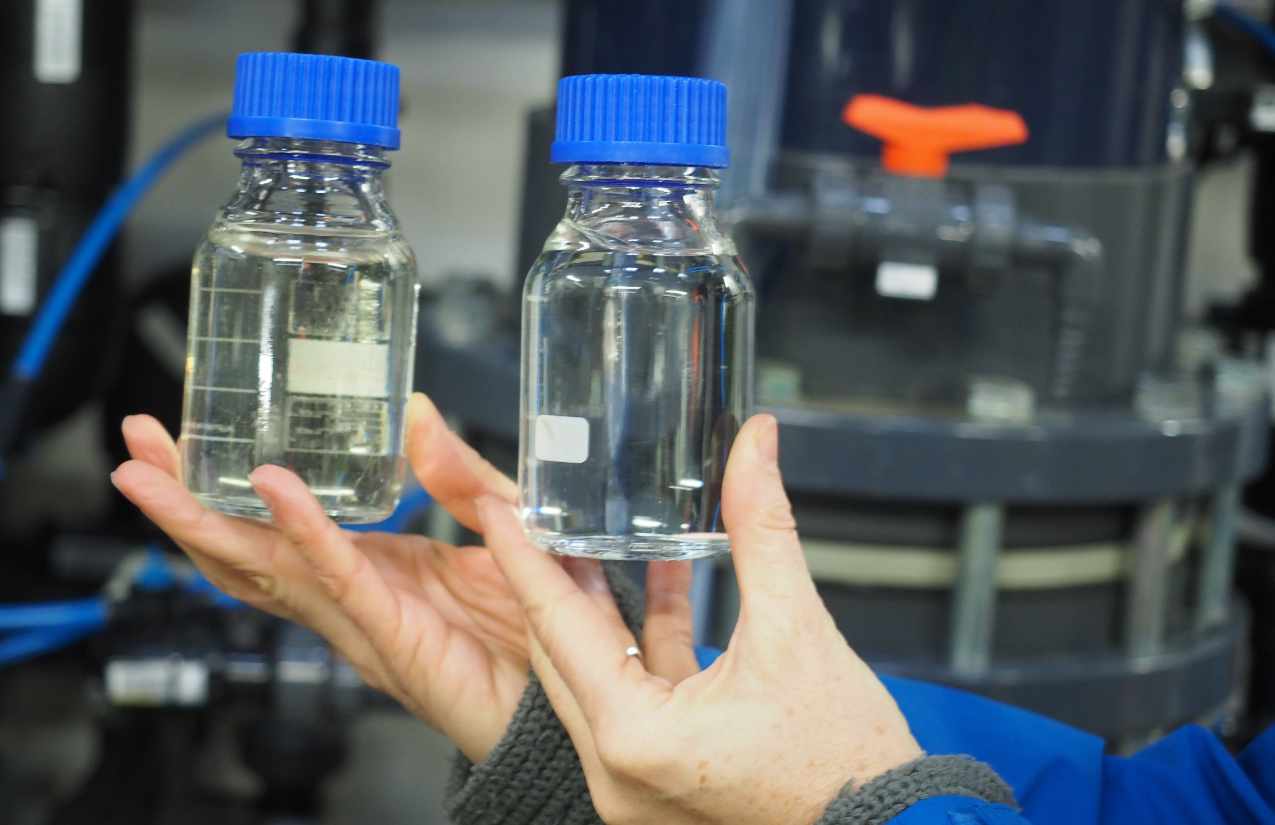 Hände halten zwei Laborflaschen mit blauem Verschluss vor einer Wasseraufbereitungsanlage