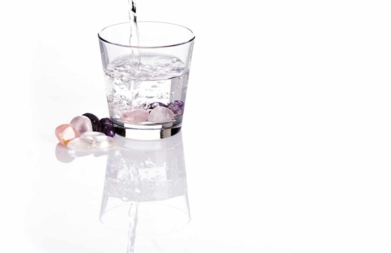 Wasser fließt in ein Glas mit Edelsteinen, weitere Trommelsteine liegen daneben auf weißem Grund