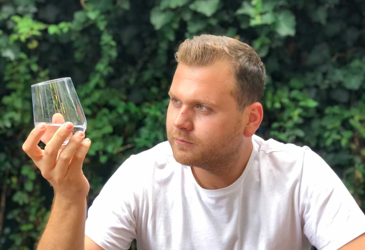 Junger Mann betrachtet prüfend ein Glas Wasser vor einer grünen Efeuwand