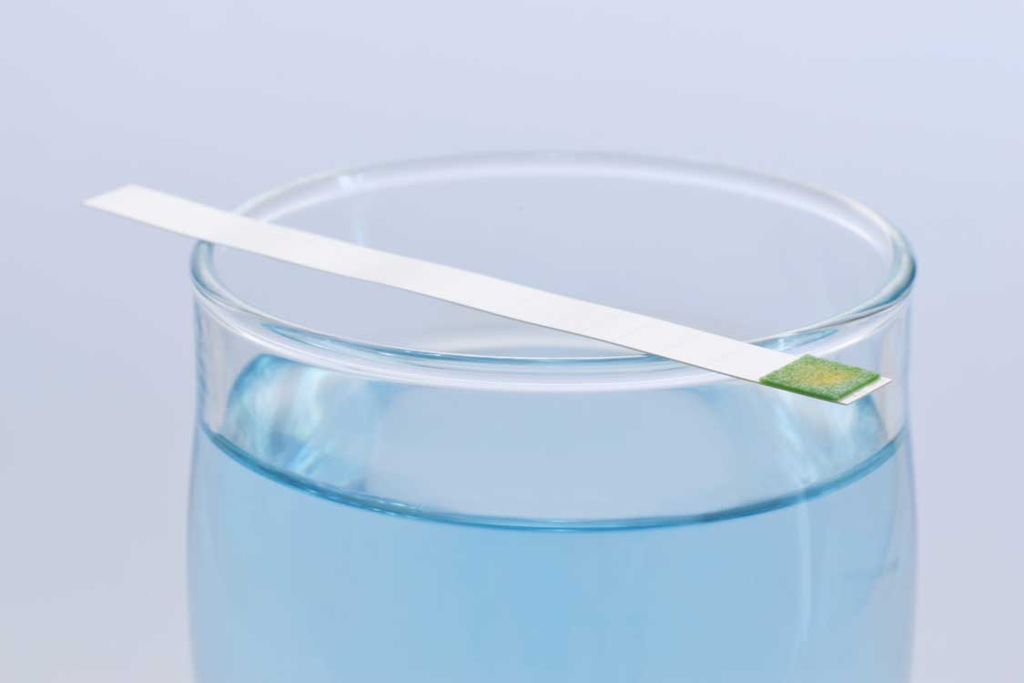 Teststreifen zur Wasserhärte liegt auf einem Glasgefäß mit Wasser, Testfeld grün verfärbt