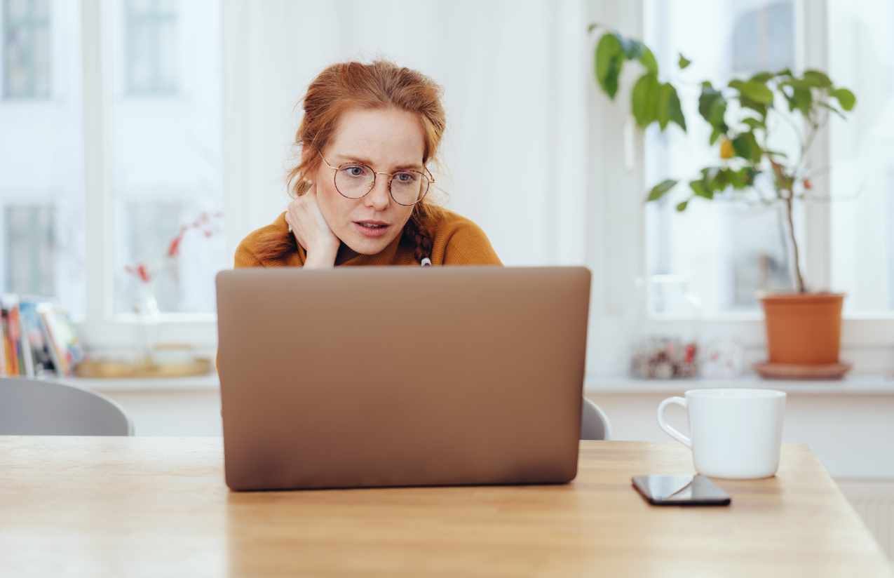 Frau mit Brille recherchiert am Laptop am Küchentisch, Tasse und Smartphone daneben