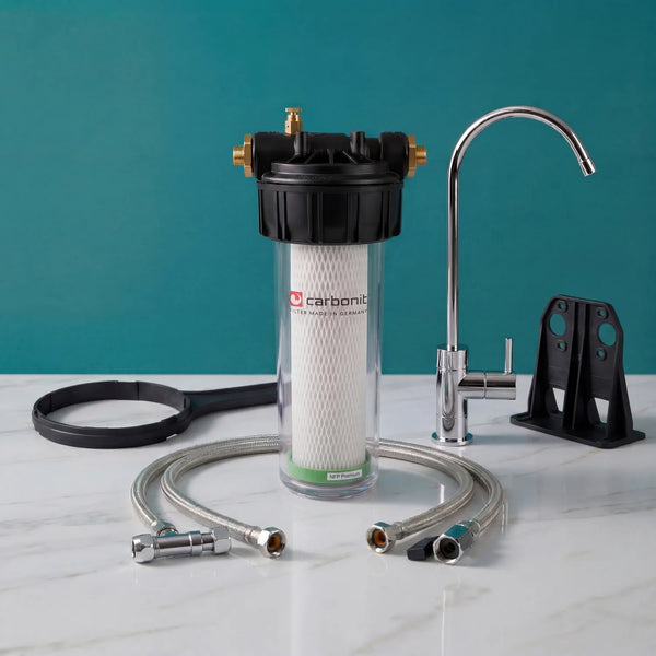 Carbonit Vario Classic Einbaufilter-Set auf weißer Marmorarbeitsplatte – transparentes Gehäuse mit NFP-Premium-Filterkartusche (Kreuzstruktur, grüner Ring), schwarzer Kopf mit Messing-Gewindeanschlüssen und Entlüftungsschraube, schwarzer Ringschlüssel und Wandhalterung, Beistell-Trinkhahn (Schwanenhals mit Hebel), zwei Schläuche; teal Hintergrund