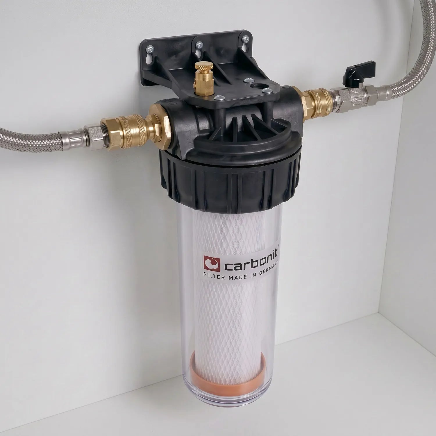 Carbonit Vario-HP Comfort montiert an weißer Schrankwand – transparentes Gehäuse mit weißer IFP-Puro-Kartusche (orangene Bodenkappe), schwarzer Kopf mit Messing-Entlüftungsschraube, Schläuche links und rechts (rechts mit schwarzem Kugelventil-Hebel)