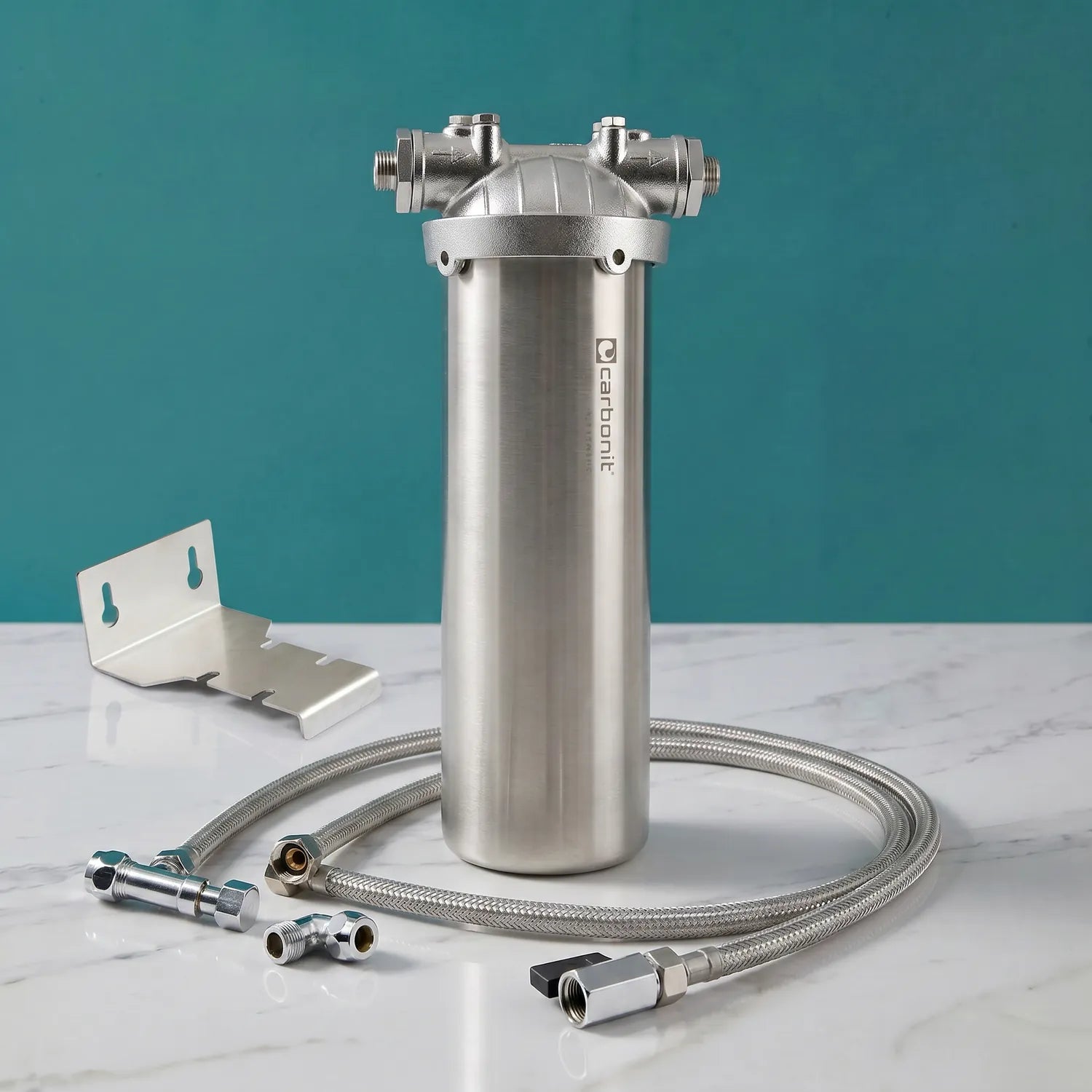 Carbonit Vario Inox VA Einbaufilter-Set auf weißer Marmorarbeitsplatte – gebürstetes Edelstahlgehäuse mit Carbonit-Logo, Gusskopf mit zwei Gewindeanschlüssen, Edelstahl-Wandhalter (L-Form), langer gewickelter Edelstahlschlauch, Winkelstücke und Fittings; teal Hintergrund