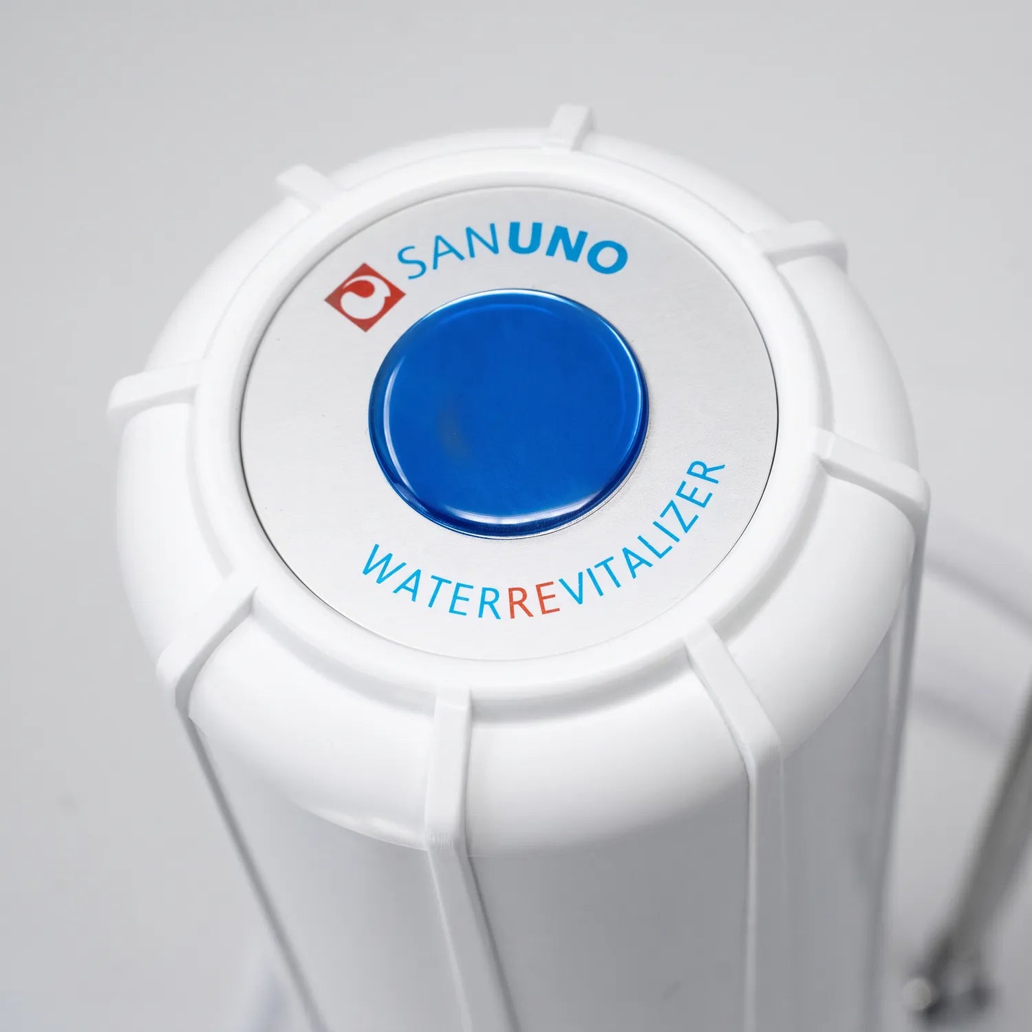 Nahaufnahme Carbonit Sanuno Auftischfilter Deckel von oben – Sanuno-Logo mit blauem Dome und „Waterrevitalizer"-Schriftzug auf weißem Kunststoffgehäuse, neutraler grauer Hintergrund